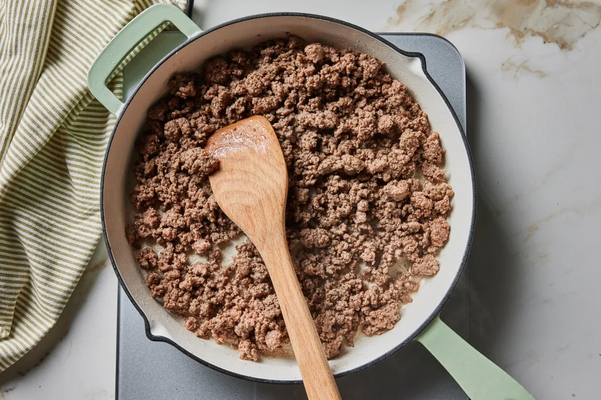 A pan filled with ground beef, accompanied by a wooden spoon resting on the side Cook with Ground Beef